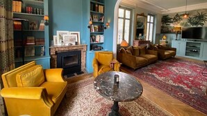 A living room with a fireplace, bookshelves, a yellow sofa, a round coffee table, and a red patterned rug. - Villa Gabriel Maison Blanche Chartres (Chartres)