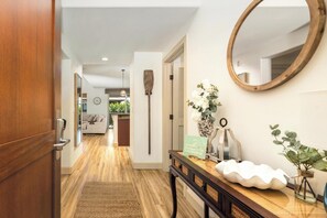 A hallway with a wooden door, a console table with a mirror, a decorative sign, and a potted plant. - Halii Kai 23G | Luxury Waikoloa Villa with Exclusive Ocean Club Access (WAIKOLOA)