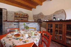 Un coin repas avec un mur en briques, des poutres en bois au plafond, une table recouverte d’une nappe à motifs floraux et un buffet aux portes en verre.