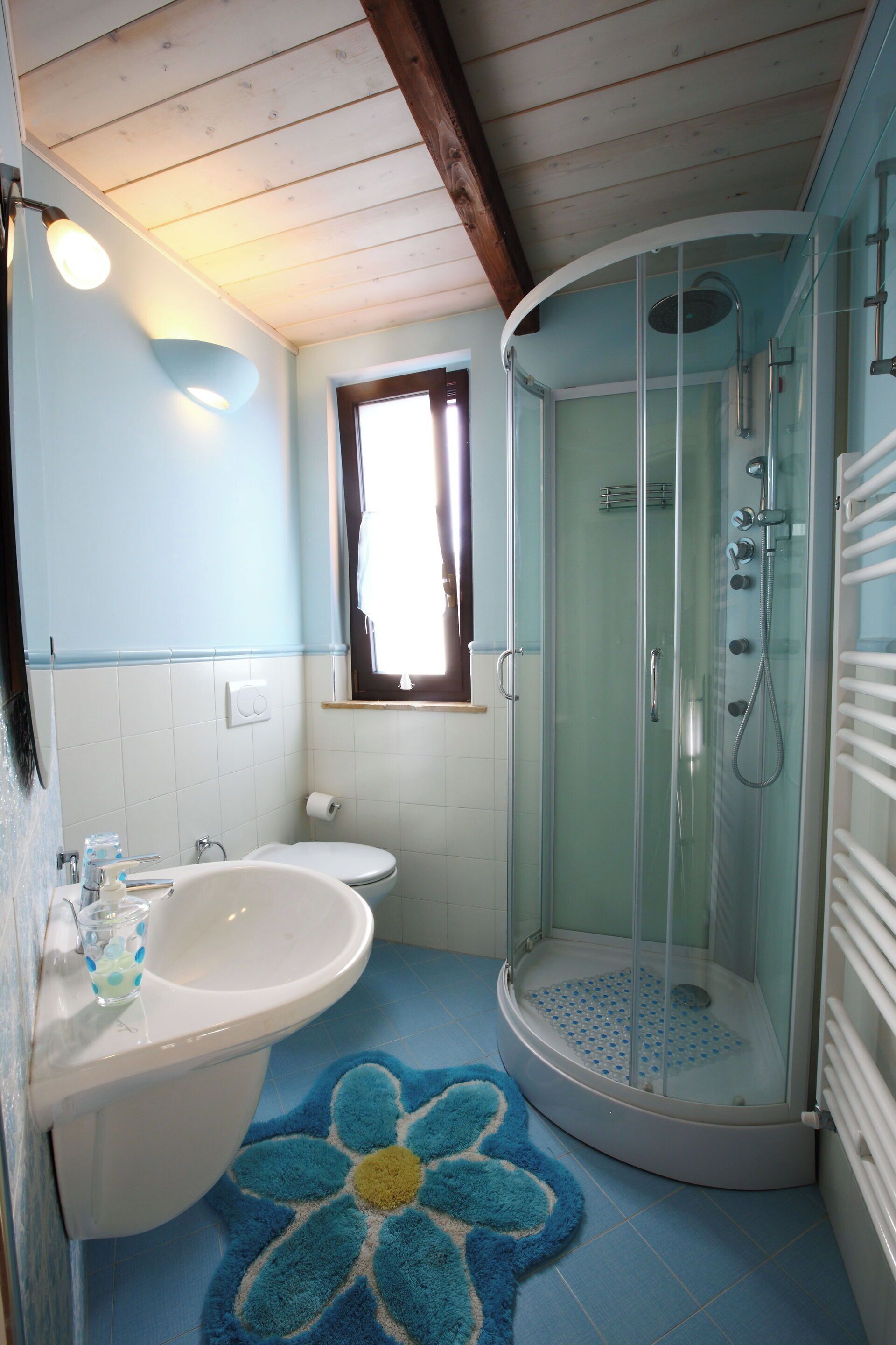 Een badkamer met een douche met glazen wand, een witte wastafel en een blauwe badmat.