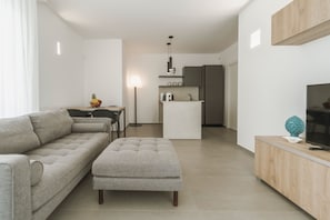 A modern living room with a sofa, ottoman, dining table, and a kitchen area in the background. - Venti di Laguna Apartments Stagnone - Tramontana (Marsala)