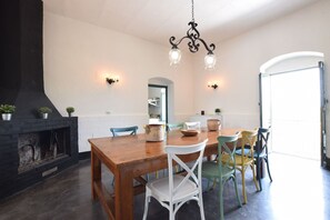 Spiseområde med et træbord, hvide stole, en sort pejs og en lysekrone.