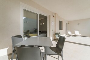 En udendørs terrasse med et bord og stole, en glasdør der fører til en balkon, og en hvid stol udenfor.