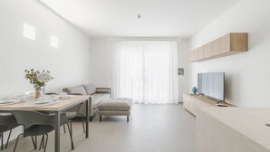 Una stanza moderna e minimalista con angolo ristoro, divano, televisione e un'ampia finestra con tende leggere.