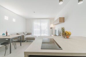 A modern kitchen with a central island, a dining area with a table and chairs, a sofa, and a large window with sheer curtains. - Venti di Laguna Apartments Stagnone - Ponente (Marsala)