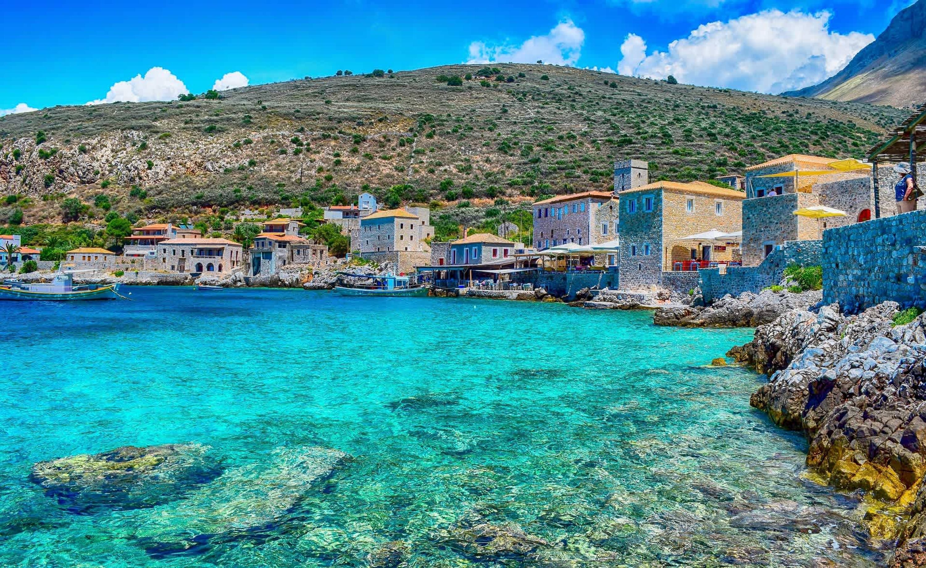 A coastal village with stone buildings, clear turquoise waters, and a hilly backdrop.