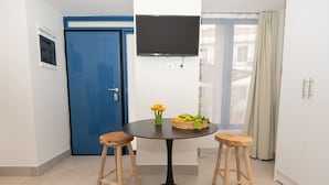A room with a round table, two wooden stools, a television on the wall, a blue door, and a window with curtains. - Gytheio Charming Studio - Steps Away From the Sea (East Mani)