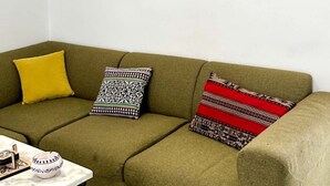 Un canapé vert avec des coussins à motifs, une petite table sur laquelle se trouve un bol, et un miroir fixé au mur.