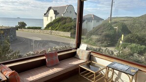 A room with a large window, a cushioned bench, a small table, and a view of a coastal road and buildings. - Family Beach House for 2-9 people near Baby Bay in New Polzeath, North Cornwall (New Polzeath, Wadebridge)