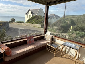 Ein Zimmer mit großem Fenster, einer gepolsterten Bank, einem kleinen Tisch und Blick auf eine Küstenstraße und Gebäude.