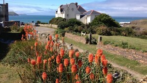 A coastal house with a stone pathway, a garden with orange flowers, and a view of the sea. - Family Beach House for 2-9 people near Baby Bay in New Polzeath, North Cornwall (New Polzeath, Wadebridge)
