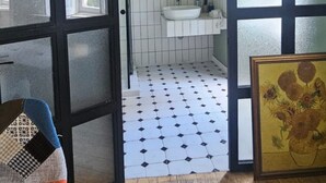 A room with a wooden floor, a black-framed door leading to a bathroom with white tiles, a sink, and a framed painting. - NinasPlace3 (T'bilisi)