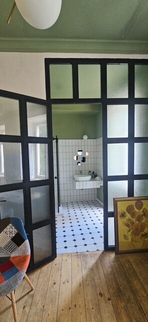 Ein Zimmer mit Holzboden, einer schwarzen Tür, die ins Badezimmer mit weißen Fliesen, Waschbecken und einem gerahmten Gemälde führt.