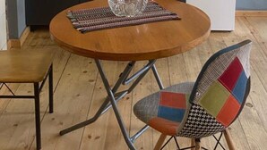 A compact kitchen with a sink, microwave, and white refrigerator. A round wooden table with a glass bowl and a woven placemat is in the foreground. A colorful chair is next to the table. - NinasPlace3 (T'bilisi)