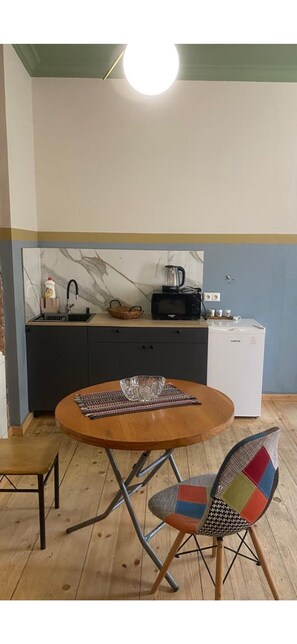 Eine kompakte Küche mit Spüle, Mikrowelle und weißem Kühlschrank. Im Vordergrund steht ein runder Holztisch mit einer Glasbowl und einer gewebten Tischmatte. Ein farbenfroher Stuhl steht neben dem Tisch.