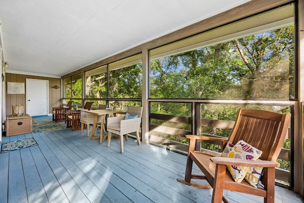 A covered porch with wooden flooring, wicker furniture, and large windows offering a view of trees.