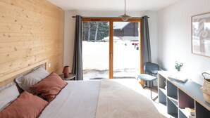 A bedroom with a wooden headboard, a bed with white and brown bedding, a desk with a chair, and a view of a snowy landscape through a sliding door. - Cosy new apartment, terrace and small garden, magnificent mountain views (Le Monêtier-les-Bains)