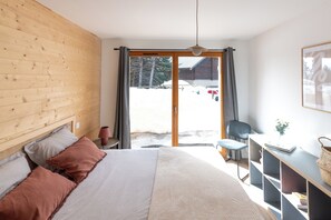 Ein Schlafzimmer mit einem hölzernen Kopfteil, einem Bett mit weißer und brauner Bettwäsche, einem Schreibtisch mit Stuhl und Blick auf eine verschneite Landschaft durch eine Schiebetür.