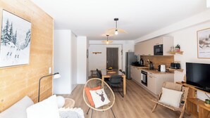 A modern living room with a sofa, a chair, a dining area, and a kitchen with a TV. - Cosy new apartment, terrace and small garden, magnificent mountain views (Le Monêtier-les-Bains)