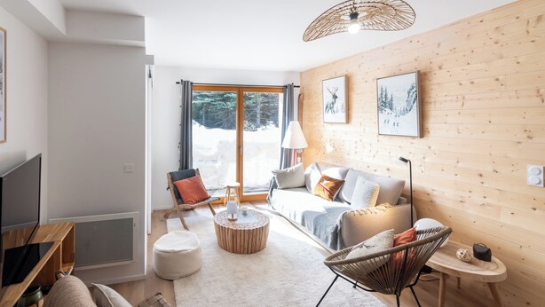 A modern living room with a sofa, armchair, and coffee table, wooden walls, and a large window with a view of trees. - Cosy new apartment, terrace and small garden, magnificent mountain views (Le Monêtier-les-Bains)