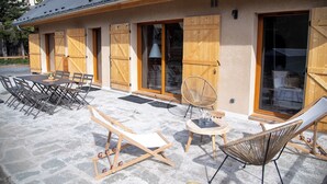 A building with a covered patio, wooden shutters, a dining table with chairs, and lounge chairs. - Cosy new apartment, terrace and small garden, magnificent mountain views (Le Monêtier-les-Bains)