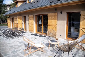 Ein Gebäude mit einer überdachten Terrasse, Holzläden, einem Esstisch mit Stühlen und Liegestühlen.