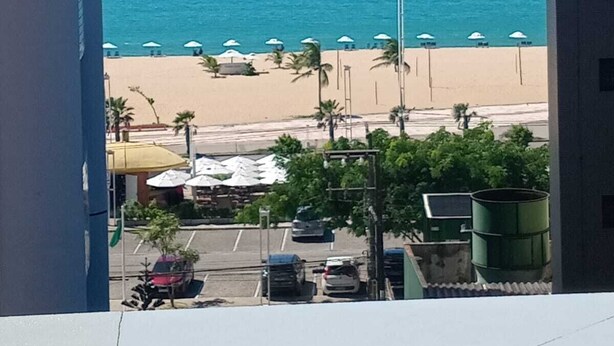 Vista de una playa con sombrillas y palmeras, un estacionamiento y un edificio al frente.