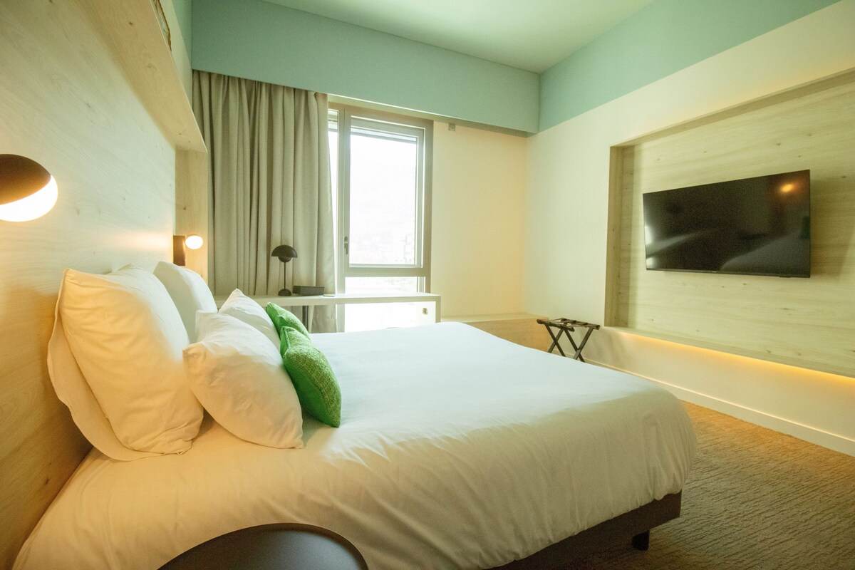 Habitación de hotel con cama, televisor montado en la pared, ventana con cortinas y mesita de noche.