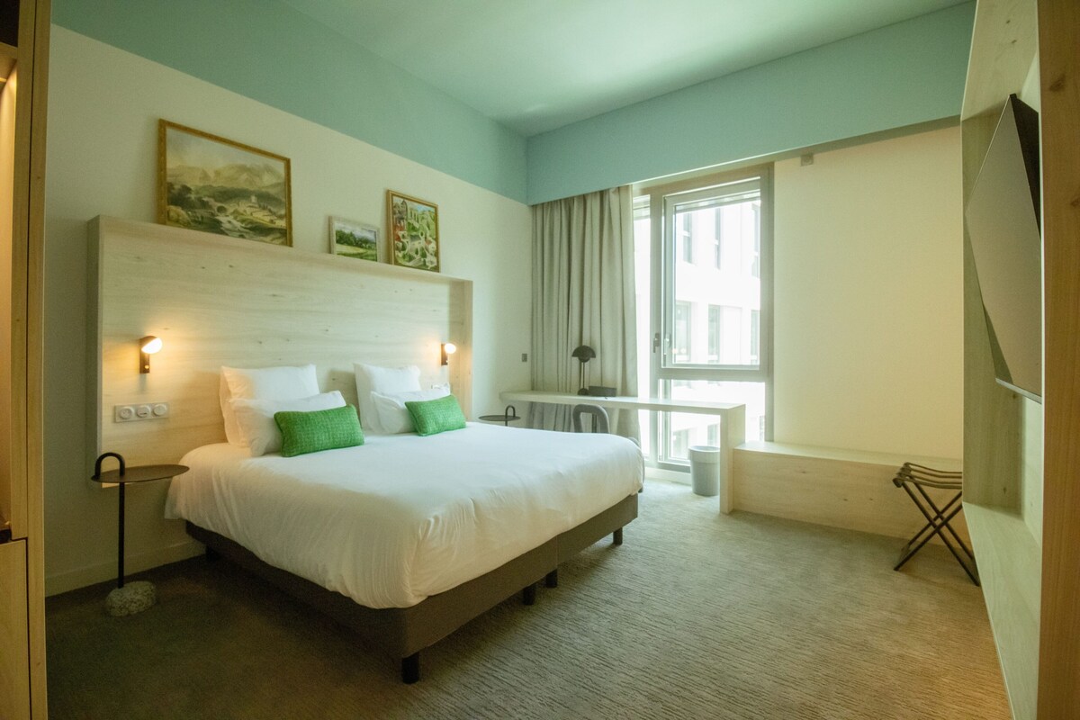 Ein Hotelzimmer mit einem großen Bett, Nachttischlampen, einem Schreibtisch und einem Fenster mit Vorhängen.