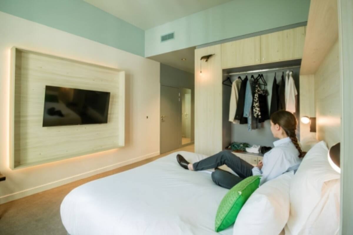 A hotel room with a bed, a TV mounted on the wall, a closet with clothes, and a door leading to another room.