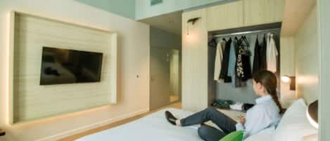A hotel room with a bed, a TV mounted on the wall, a closet with clothes, and a door leading to another room.