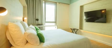 Habitación de hotel con cama, televisor montado en la pared, ventana con cortinas y mesita de noche.