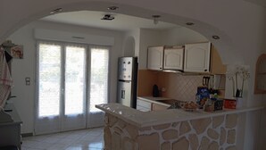 Cozinha com ilha de pedra, armários brancos, geladeira e vista através de uma porta de vidro.