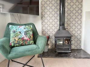 A green armchair with a floral cushion, a wood-burning stove, and patterned wallpaper. - One bed Character Cottage with Parking and Garden (East Portlemouth)