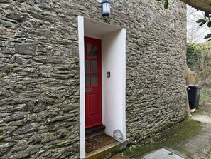 Un bâtiment en pierre avec une porte rouge, un petit marchepied menant à l’entrée et une poubelle noire sur la droite.