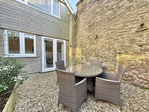 Une terrasse avec une table ronde et des chaises en osier, un mur en pierre et un bâtiment en bois doté de fenêtres.