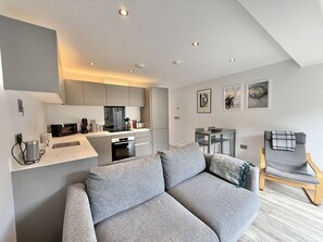 Ein modernes Wohnzimmer mit grauem Sofa, einem Holzstuhl, einer Küchenzeile und einem Esstisch.