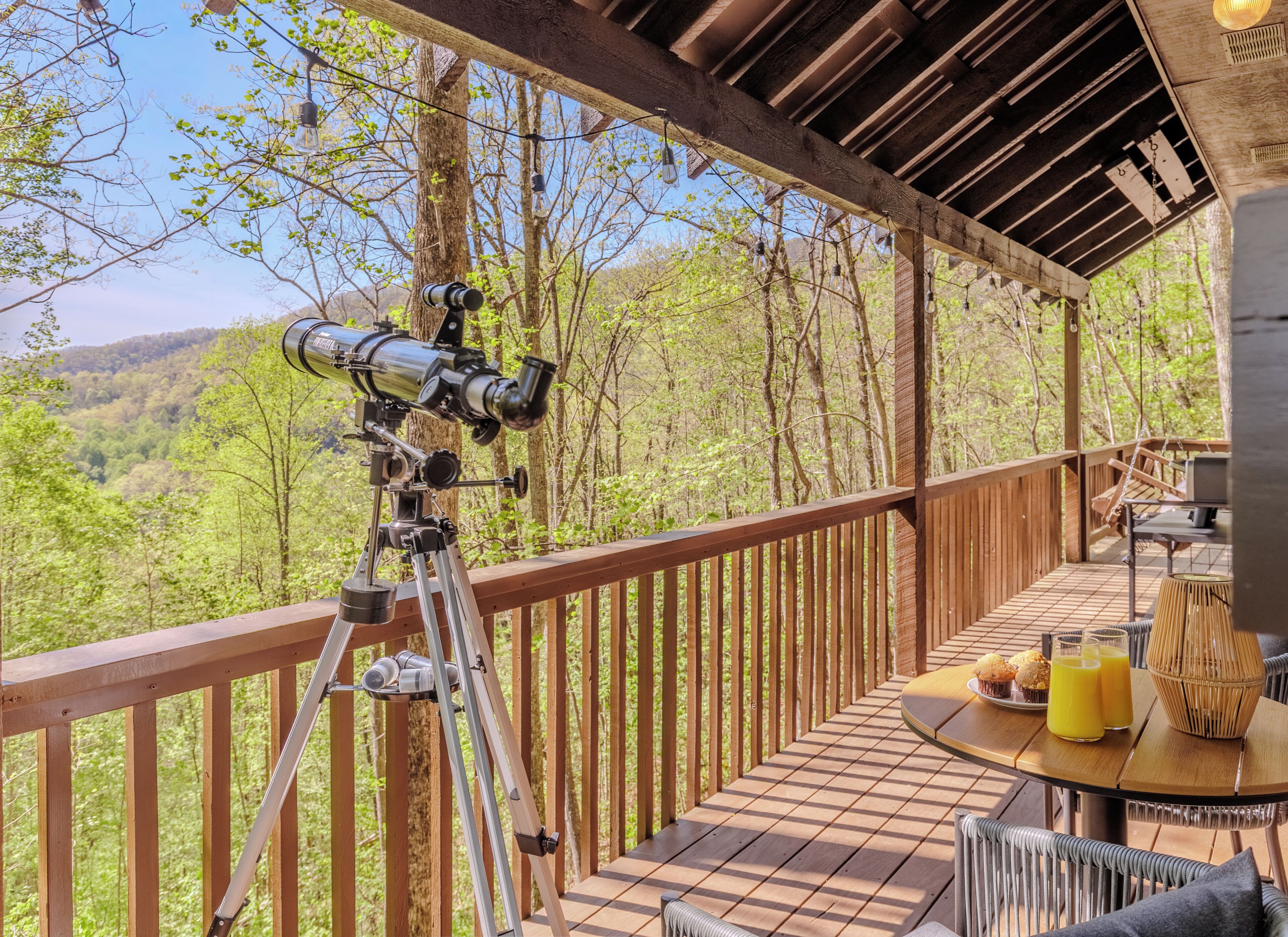 A wooden deck with a telescope, a table with drinks and snacks, and a view of a forested area.