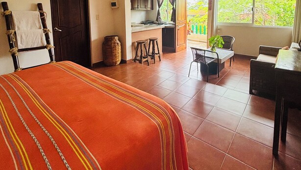A hotel room with a bed, a kitchenette, a dining area, and a large window with a view of greenery.