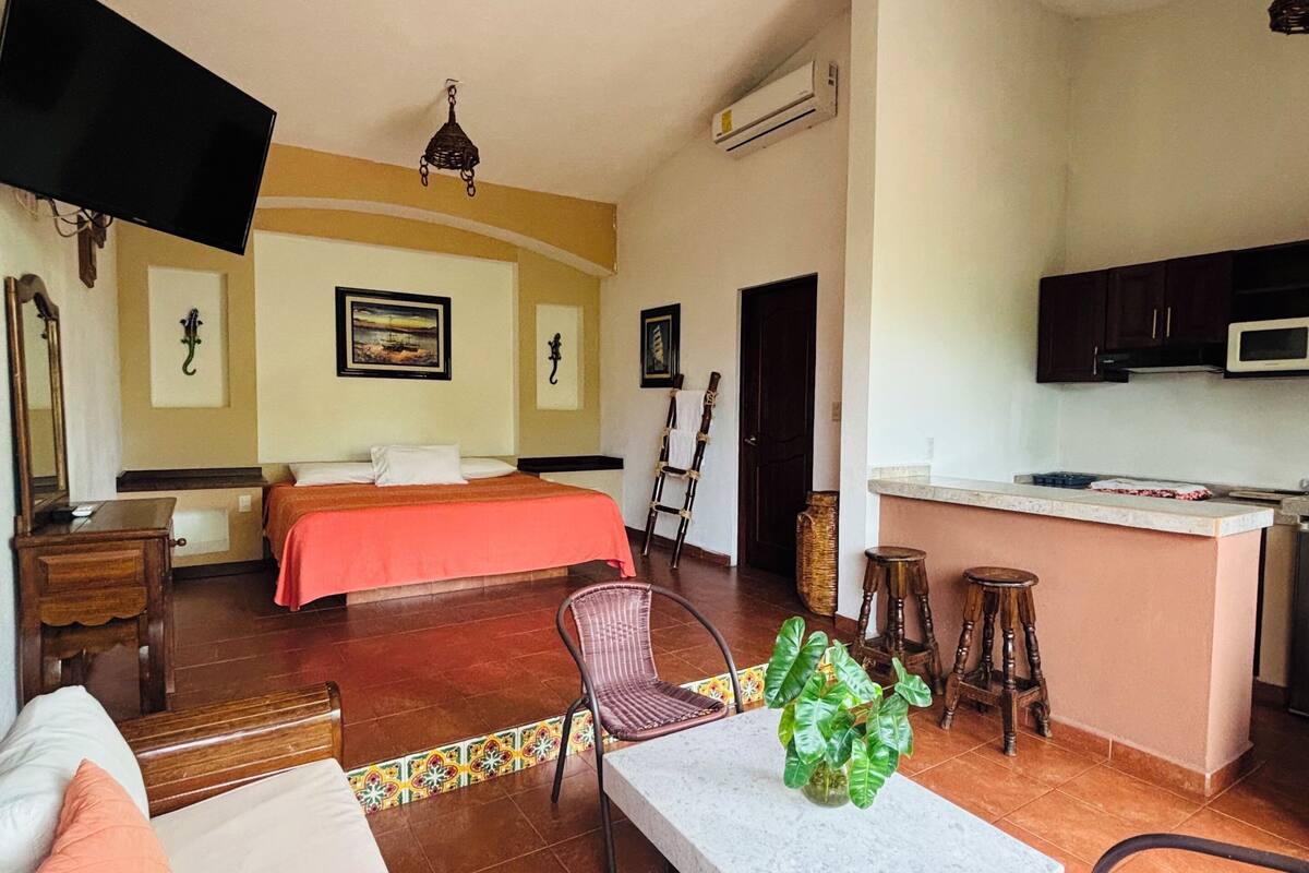 Habitación de hotel con cama, sofá, mesa de comedor, sillas, televisión, zona de cocina y ventilador de techo.