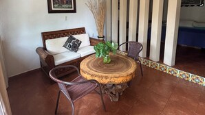 A room with a round wooden table, two wicker chairs, a sofa with a cushion, a framed picture on the wall, and a vertical striped partition.