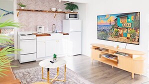 A modern kitchen with a white stove, white cabinets, a microwave, and a sink. There's a round table with a plant, a wooden cabinet with books, and a painting of a street scene on the wall. - Sweet Stowe Apartment (Stowe)
