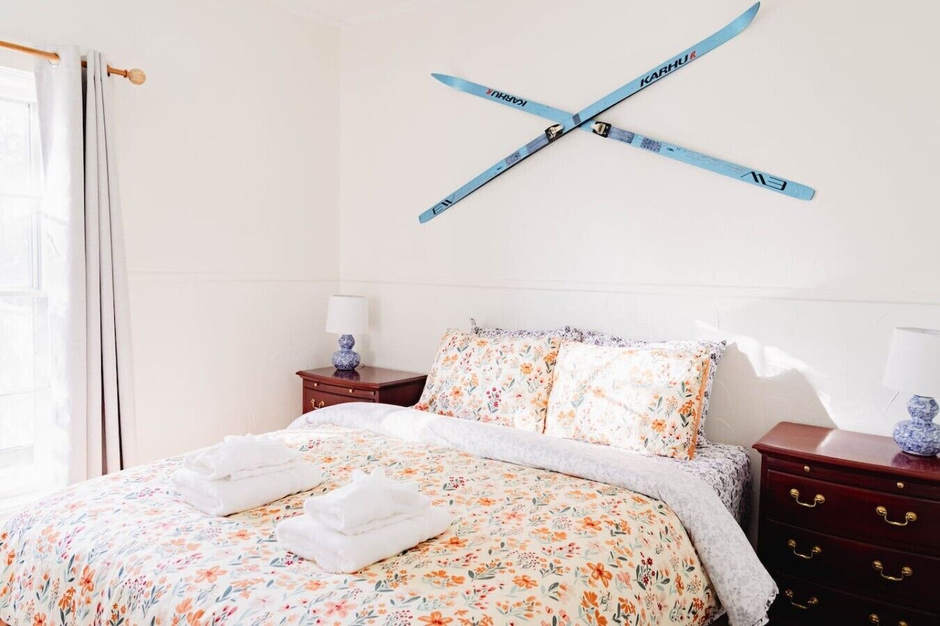 Een slaapkamer met een bed, bloemenbedrukking, twee nachtkastjes met lampen en een raam met gordijnen.