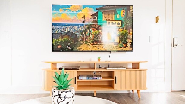 A modern living room with a flat-screen TV mounted on the wall, a wooden entertainment unit below, and a round coffee table with a remote control and a potted plant. - Sweet Stowe Apartment (Stowe)