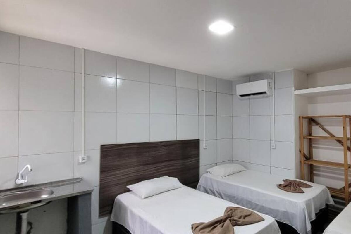 A hotel room with two single beds, a small desk, a chair, a TV, a shelf with a ladder, and a wall-mounted air conditioner.