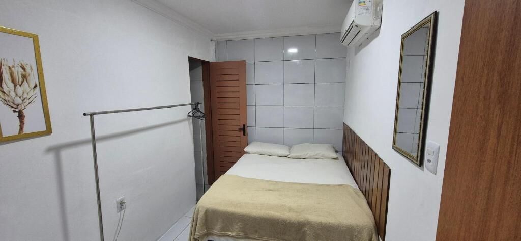 Quarto de hotel pequeno e estreito, com uma cama, uma porta de madeira, um porta-toalhas e um ar condicionado fixado na parede.