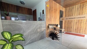 A reception area with a wooden wall, a glass door with a sign, a potted plant, and a tiled floor. - Pousada Primeiro Sol (João Pessoa)