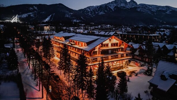 A large, multi-story lodge with a prominent front entrance, surrounded by snow-covered trees and mountains in the background. - Mountain Diamond Resort (Zakopane)