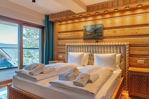 Habitación de hotel con una cama grande, paneles de madera, un cuadro sobre la cama y una ventana con vista a árboles.