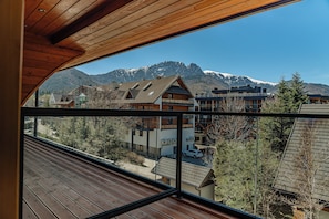 Vista desde un balcón con barandilla de madera, que da a un paisaje montañoso con picos nevados y una hilera de edificios con tejados inclinados.